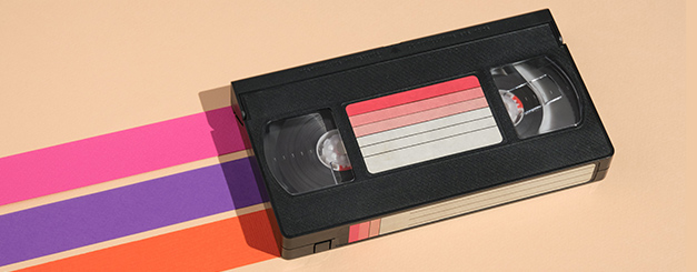 A black VHS tape placed diagonally on a light tan surface, featuring a label with horizontal red and white gradient stripes. Behind the tape are three parallel, vibrant bands of pink, purple, and orange extending across the frame.