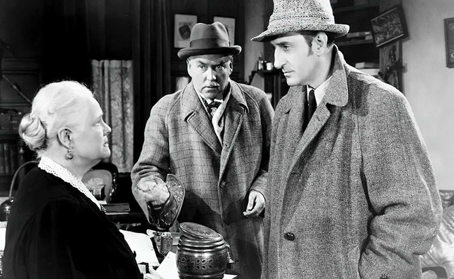 Still from "Dressed to Kill". Black and white image of two men in wool coats and hats speaking to an older woman. The setting is a cozy, cluttered room, conveying a sense of mystery. From left to right: Marjorie Bennett as Antique Shop Assistant, Nigel Bruce as Doctor Watson, Basil Rathbone as Sherlock Holmes.