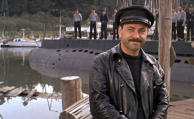 Still from "The Russians Are Coming, the Russians Are Coming". A man with a mustache in a black leather jacket and cap, Alan Arkin as Lieutenant Rozanov, smiles slightly in front of a docked submarine. Sailors stand on the submarine in the background.