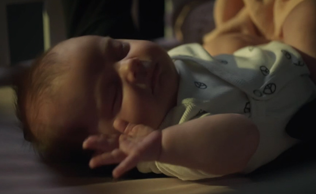 Uncredited actor in Luna's role, a peaceful baby with closed eyes sleeps in a crib while wearing a light-colored onesie with small peace sign patterns, one hand gently raised, captured in soft, dim lighting.