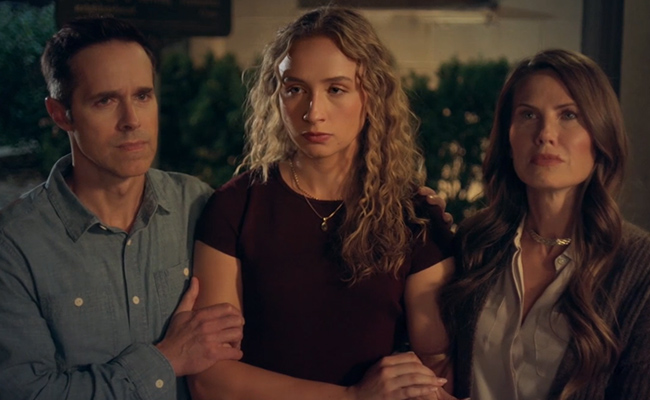 Tim Bensch as Don, wearing a blue button-down shirt, has his hand on Tatum Jennings as Carly, a young woman with curly blonde hair wearing a maroon short-sleeved top and a delicate gold necklace and Vail Bloom as Suzanne, a woman with long brown hair wearing a light-colored blouse under a dark cardigan stand close together in a dimly lit outdoor environment, all looking forward with serious, somber expressions.