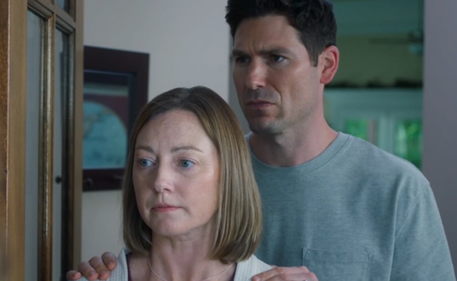 Robbie Silverman as Pastor David, a man with dark hair wearing a light gray t-shirt stands behind Jennice Butler as Isabella, a woman with blonde shoulder-length hair, resting his hands on her shoulders. Both appear serious as they look forward in an indoor setting.