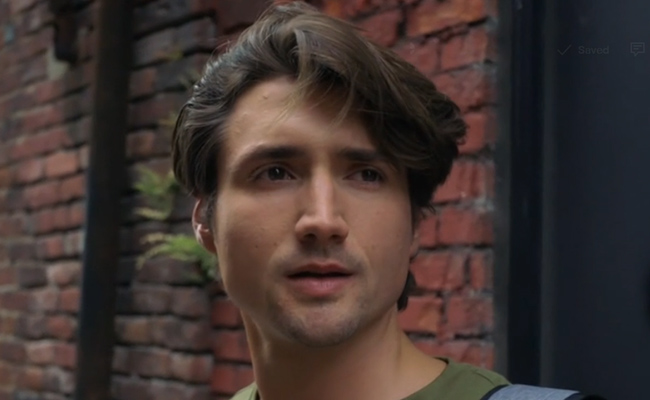 Close-up still of Kyle-X Storm as Marcus, a young man with wavy dark hair looking off-camera with a contemplative expression, wearing a green shirt in front of an exterior red brick wall.