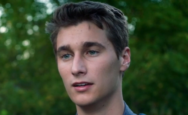 A head-and-shoulders still of Peyton Windham as Brayton Grey, a young man with short, tousled light brown hair and blue eyes, positioned against a soft-focus background of green trees.