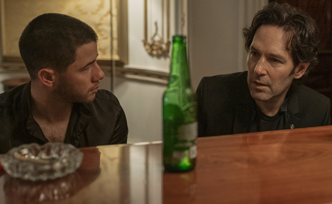 Still from "Power Ballad". Two men sit at a piano, engaged in a serious conversation. On the left is Nick Jonas as Danny, with close-cropped, dark hair, wearing a black shirt unbuttoned at the neck. On the right is Paul Rudd as Rick, with longer wavy brown hair parted in the middle, wearing a black blazer and T-shirt. A green bottle of beer and a glass ashtray are on what seems to be an upright piano. The scene is dimly lit and tense.