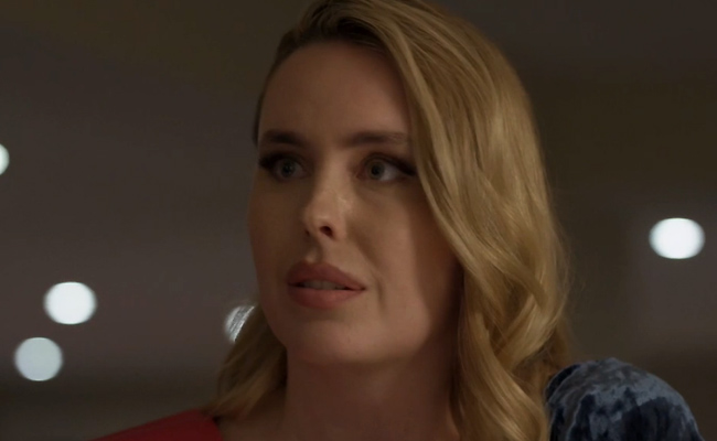 A close-up of Kirsten Comerford as Lori, a blonde woman with a concerned, slightly shocked expression and her mouth parted. She is wearing red sash over a textured dark blue garment, set against a dark, out-of-focus background with blurred bokeh lights.