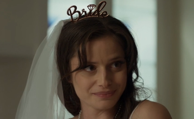 Rachel Sellan as Angela. A young woman with brown hair wears a white bridal veil and a rose gold "Bride" tiara featuring a diamond icon, smiling subtly as she looks off to the side.