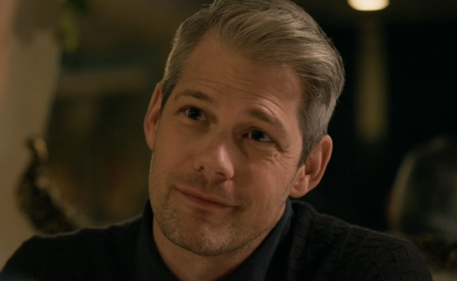 A close-up of Jesse Collin as James, a man with neatly styled salt-and-pepper hair and light stubble, wearing a dark textured sweater over a black shirt, looking slightly off-camera with a warm, subtle smile.