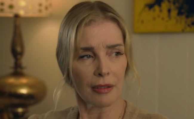 A medium close-up of Jayne Heitmeyer as Frances, a blonde woman with a somber, pensive expression, wearing a beige knit top. In the background, out-of-focus elements include a metallic table lamp and an abstract yellow painting.