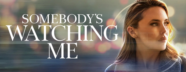 A promotional graphic for "Somebody's Watching Me" featuring Kirsten Comerford as Lori, a woman with highlighted blond hair swept back held by four Bobby pins looking off to the side, set against a dark background with blurred city lights and horizontal light streaks.