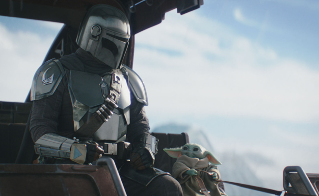 Still from "Star Wars: The Mandalorian and Grogu" featuring Pedro Pascal as Din Djarin, a bounty hunter in Mandalorian armor, sitting next to a small, green creature, Grogu, swaddled in a robe, both appearing to be inside a vehicle against a bright, hazy sky.