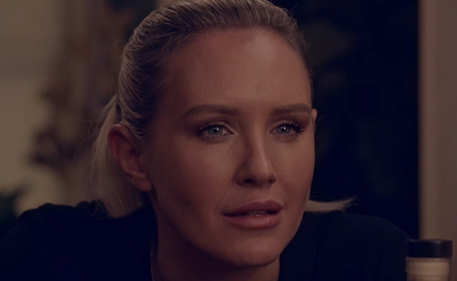 Close-up of Nicky Whelan as Kate, a woman with blonde hair pulled back, looking off-camera with a somber, focused expression in a dimly lit indoor setting.