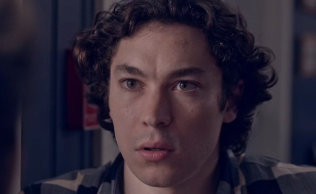 A close-up still of Nikolas Elrifi as Mark, a young man with dark, curly hair and a fair complexion, wearing a dark plaid shirt. He is looking forward with a serious, slightly concerned expression, his mouth slightly parted. The background is softly blurred and dimly lit.