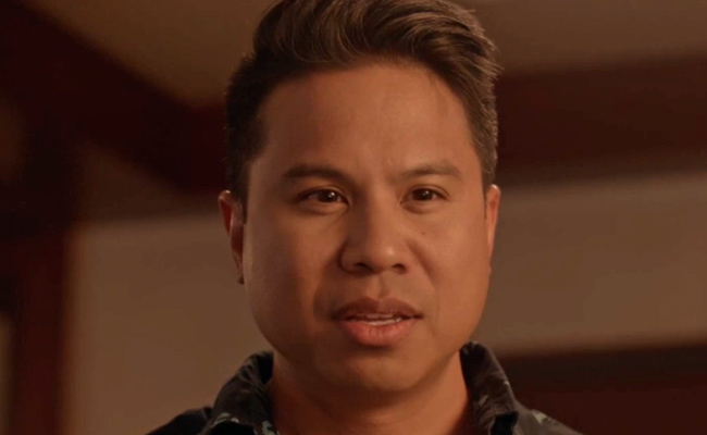 A close-up still of Ric Plamenco as Colin, a man with dark, styled hair and brown eyes, wearing a dark patterned shirt, looking downwards with a serious expression against a softly lit, warm-toned, and out-of-focus background.