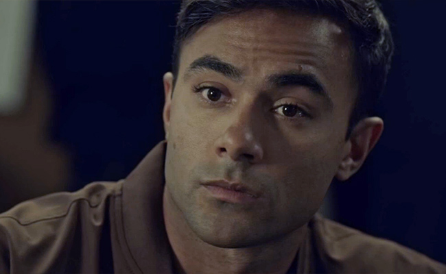 Close-up still of Brennan Mejia as Detective Lynch, a man with short, dark hair and a serious, focused expression, wearing a brown collared shirt against a dimly lit, out-of-focus background.