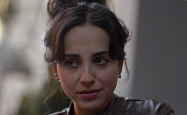 Close-up still of Skye Coyne as Danielle, a woman with dark hair styled in an updo, wearing a brown leather jacket, looking slightly off-camera with a neutral expression.