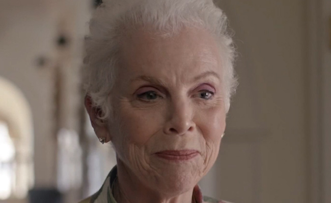 A close-up still of Ellen B. Williams as Claire, an older woman with short, white hair and fair skin, wearing subtle makeup. She has a gentle, reflective expression as she looks slightly to the side. The background is softly out of focus, suggesting the interior with an arched window.