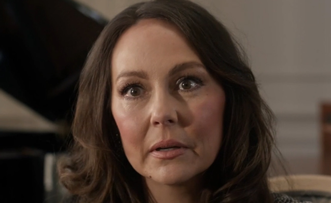 A close-up still of Christy Tate as Louise, a woman with long, dark, wavy hair looking off-camera with a neutral, attentive expression in a softly lit indoor environment.