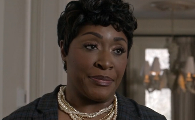 Close-up still of Monanik 'Moe' Dugar as Ingrid, a woman with short, dark curly hair wearing a dark plaid blazer and a multi-strand pearl and gold necklace, looking off-camera in a softly lit indoor setting.