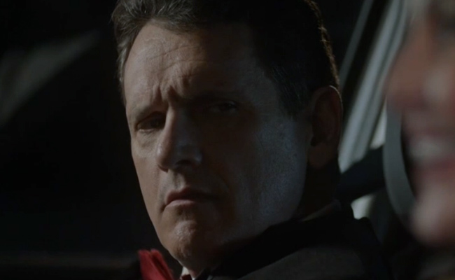 A close-up still of Eric Olson as Alex, a serious-looking man with short hair, captured in low lighting, inside a car at night, with the out-of-focus silhouette of Christy Tate as Louise, visible in the foreground.