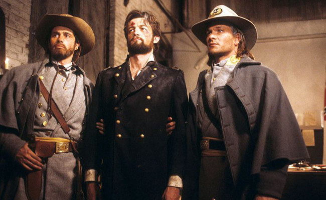 From "North and South: Book II", three men dressed in Civil War era military attire, in a large room with a white-painted brick wall with large wooden doors to the left. An unknown actor with a beard on the left sports a gray Confederate uniform and a brown hat. In the center, James Read as George Hazard, with a beard and a wound on his head, wears a black officer's overcoat. On the right, Patrick Swayze as Orry Main, is also seen in a full gray Confederate uniform.