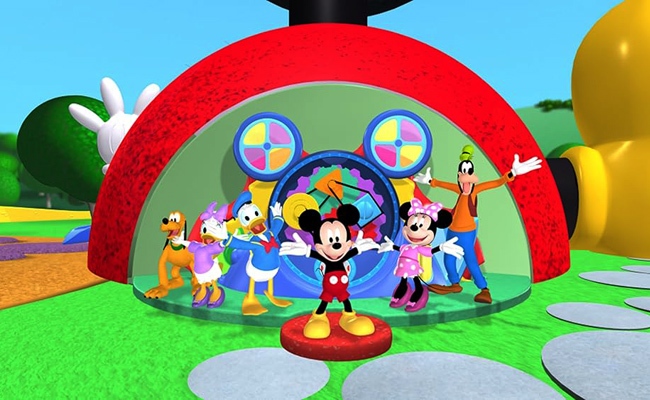 From "Mickey Mouse Clubhouse", animated characters Mickey Mouse, Minnie Mouse, Donald Duck, Daisy Duck, Goofy, and Pluto stand together on a green, grassy, cartoonish playground in front of a partially visible red and yellow structure with a Mickey Mouse-themed background featuring spinning gears and colorful wheels.