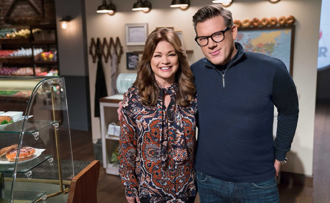 From "Food Netowrk Star: Comeback Kitchen", a woman, Valerie Bertinelli, with wavy brown hair wearing a patterned blouse, and a man, Tyler Florence, with glasses, wearing a navy blue quarter-zip sweater and blue jeans stand smiling in a studio set resembling a kitchen, a display case to the left with a bagel topped with cream cheese, smoked salmon, capers and onions on a white plate on a shelf inside.