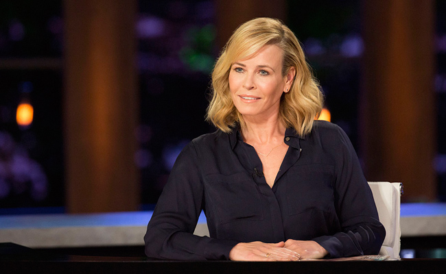 A still from the Netflix TV talk show "Chelsea". Chelsea Handler, a blonde woman wearing a dark blue button-down shirt and a necklace sits at a desk with her hands clasped, looking off to the side with a slight smile. The blurred background features decorative wood planks and purple lighting.
