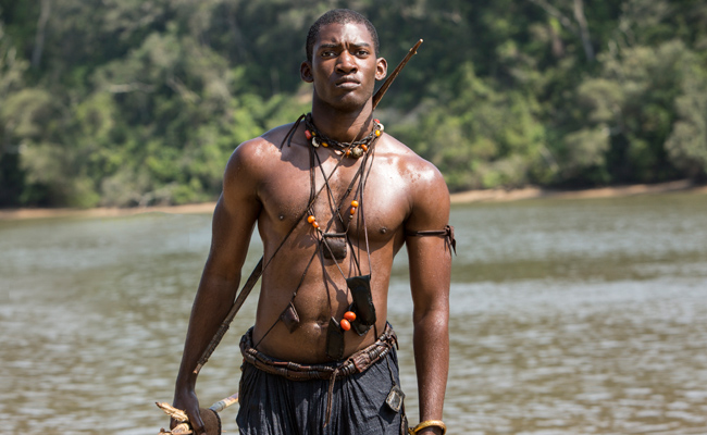Still from the 2016 TV miniseries "Roots", featuring a shirtless Black man, Malachi Kirby as Kunta Kinte, standing in a river, holding a spear in his right hand. He wears traditional African necklaces and accessories. The background is a lush forest, conveying a strong, natural tone.