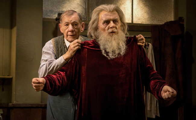 Still from TV movie "The Dresser", featuring an elderly man, Ian McKellan as Norman, helping an actor, Anthony Hopkins as Sir, wearing a fake gray beard and wig with shoulder-length gray hair, put on a red velvet robe in a dimly lit dressing room. The scene conveys a sense of care and preparation.