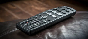 A black media remote control with a numeric keypad and navigation buttons resting diagonally on a dark brown textured leather surface.